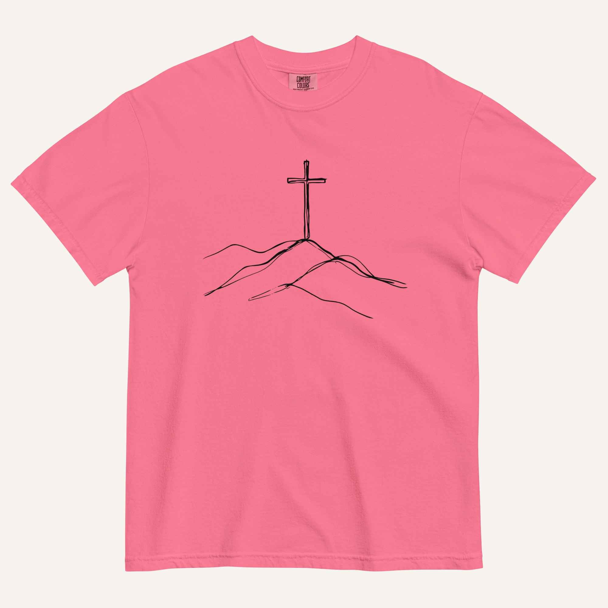A pink short-sleeve crewneck t-shirt featuring a black line art graphic of a cross on a hill, displayed flat against a light cream background.