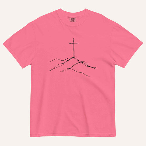 A pink short-sleeve crewneck t-shirt featuring a black line art graphic of a cross on a hill, displayed flat against a light cream background.