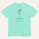 A  mint green short-sleeve crewneck t-shirt with a black line-art mustard seed graphic and the text 'FAITH as small as a mustard seed' and 'MATTHEW 17:20' printed on the front, displayed against a light cream background.