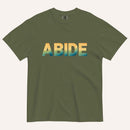 A dark green crewneck t-shirt featuring the word 'ABIDE' printed across the chest in a bold sans-serif font with a yellow-to-blue gradient.