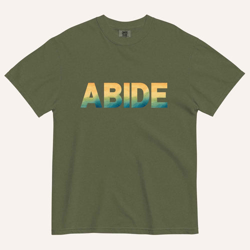 A dark green crewneck t-shirt featuring the word 'ABIDE' printed across the chest in a bold sans-serif font with a yellow-to-blue gradient.