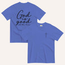 A blue short-sleeve t-shirt shown from both the front and back on a light cream background. The back features the phrase 'God is good all the time' in black script, and the front has a small black cross on the left chest.