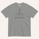 A gray short-sleeve crewneck t-shirt featuring a black line art graphic of a cross on a hill with the word 'Redeemed' printed underneath, displayed on a light cream background.