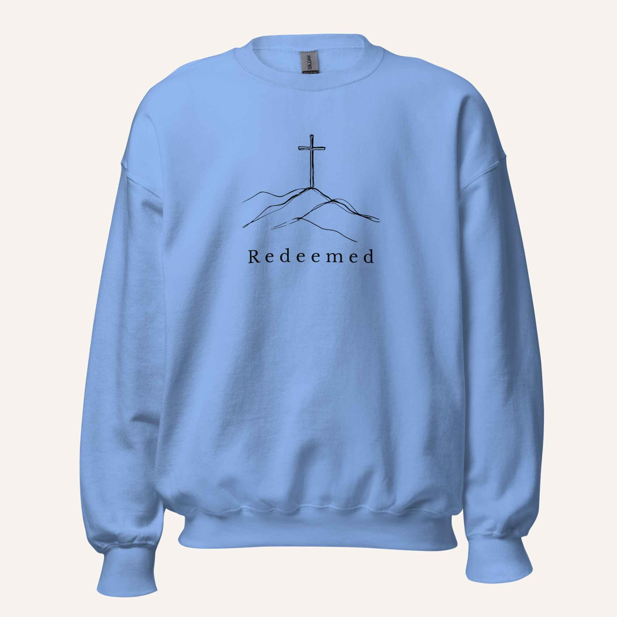 A light blue crewneck sweatshirt featuring a black graphic of a cross on a hill with the word 'Redeemed' printed below it, displayed on a clean off-white background.