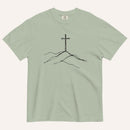 A sage green crewneck t-shirt featuring a black line art graphic of a cross rising above simple hills on the chest, displayed flat against a light off-white background.