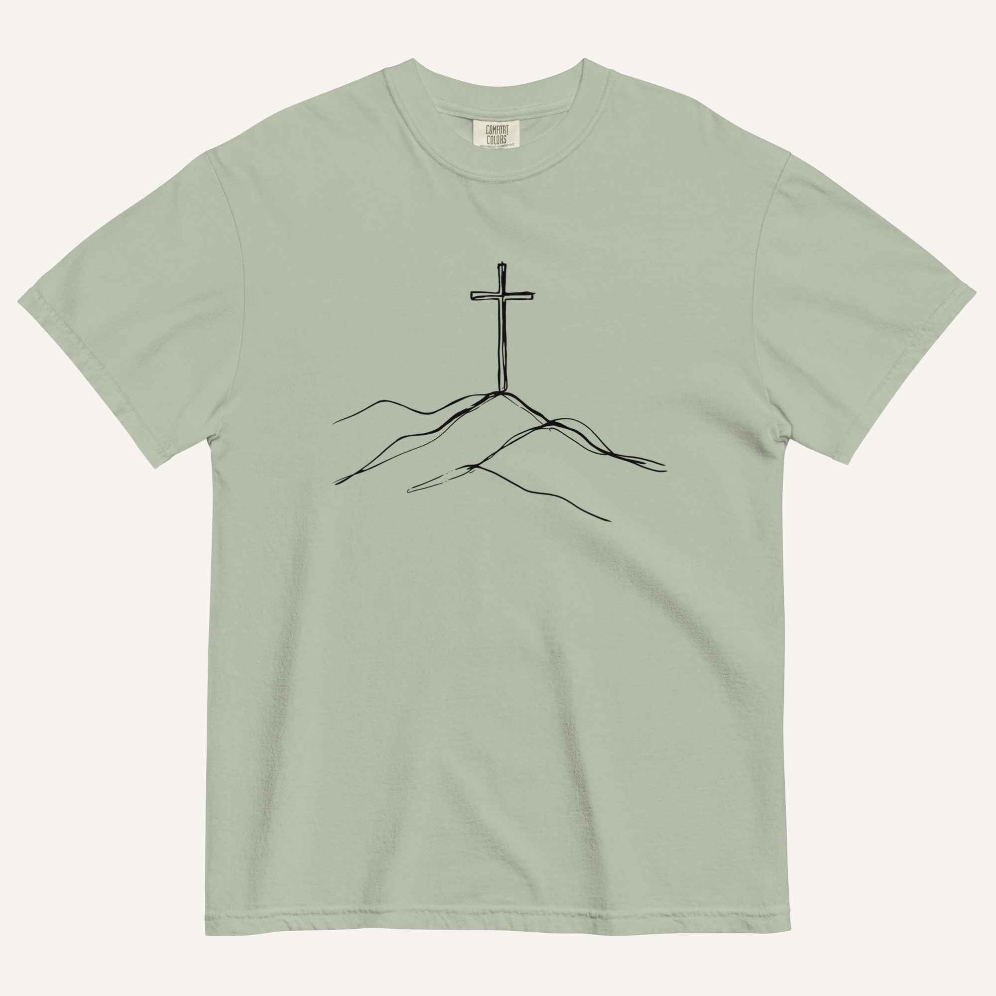 A sage green crewneck t-shirt featuring a black line art graphic of a cross rising above simple hills on the chest, displayed flat against a light off-white background.