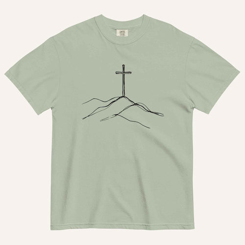 A sage green crewneck t-shirt featuring a black line art graphic of a cross rising above simple hills on the chest, displayed flat against a light off-white background.