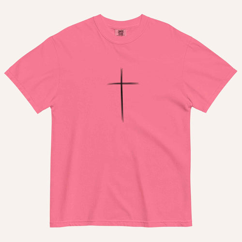 A short-sleeve crewneck t-shirt in a pink color, featuring a simple black Christian cross printed on the center chest. The t-shirt is laid flat against a solid, warm off-white background.