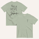 A sage green t-shirt shown from both the front and back. The front features a small black cross on the top left. The back features the black text 'There is HOPE in Jesus' in a mix of script and block lettering.