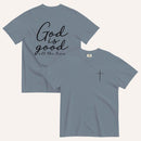 A muted dark blue short-sleeve t-shirt shown from both the front and back on a light cream background. The back features the phrase 'God is good all the time' in black script, and the front has a small black cross on the left chest.