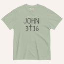 A sage green short-sleeve crewneck t-shirt with 'JOHN 3 16' text with cross graphic in between the 3 & 16 printed in a black hand-drawn font on the chest, displayed flat against a light cream background.