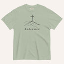A sage green short-sleeve crewneck t-shirt featuring a black line art graphic of a cross on a hill with the word 'Redeemed' printed underneath, displayed on a light cream background.