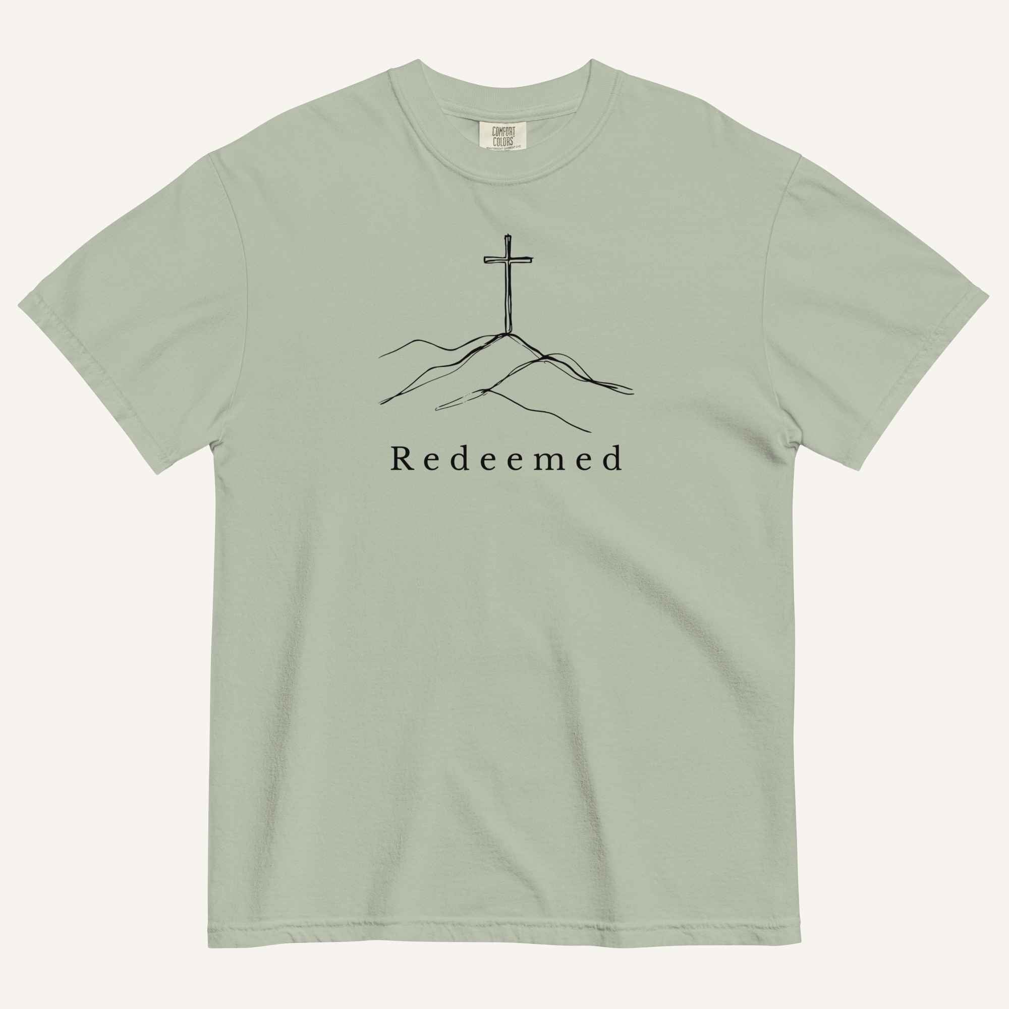 A sage green short-sleeve crewneck t-shirt featuring a black line art graphic of a cross on a hill with the word 'Redeemed' printed underneath, displayed on a light cream background.