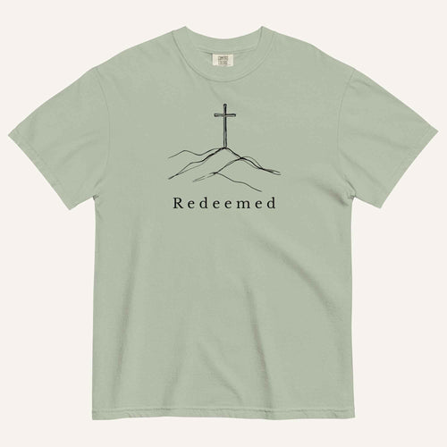 A sage green short-sleeve crewneck t-shirt featuring a black line art graphic of a cross on a hill with the word 'Redeemed' printed underneath, displayed on a light cream background.
