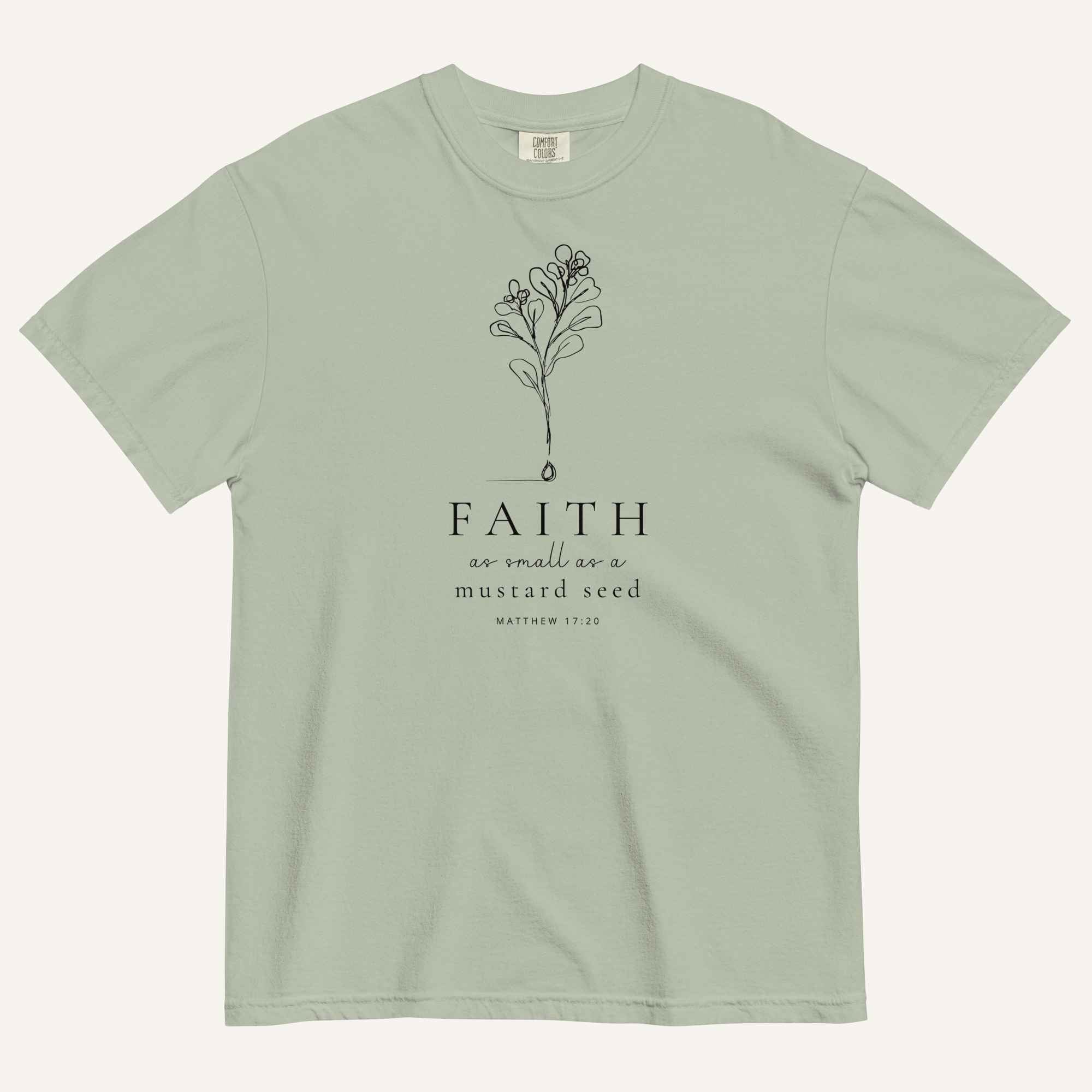 A sage green short-sleeve crewneck t-shirt with a black line-art mustard seed graphic and the text 'FAITH as small as a mustard seed' and 'MATTHEW 17:20' printed on the front, displayed against a light cream background.