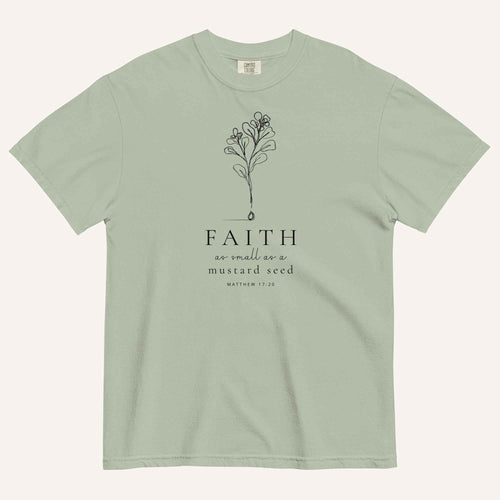 A sage green short-sleeve crewneck t-shirt with a black line-art mustard seed graphic and the text 'FAITH as small as a mustard seed' and 'MATTHEW 17:20' printed on the front, displayed against a light cream background.