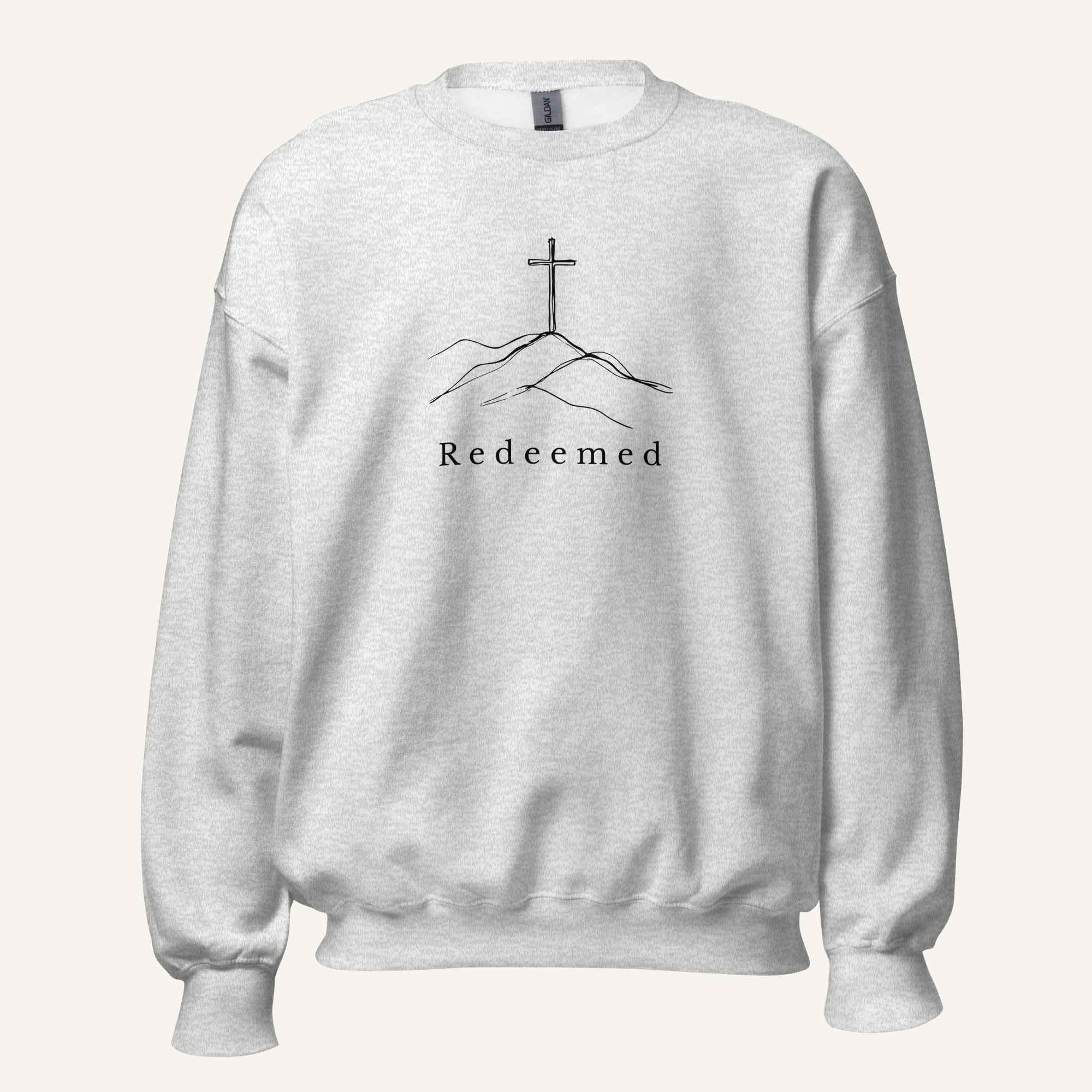 A light ash grey crewneck sweatshirt featuring a black graphic of a cross on a hill with the word 'Redeemed' printed below it, displayed on a clean off-white background.