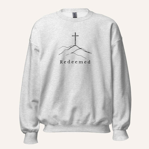 A light ash grey crewneck sweatshirt featuring a black graphic of a cross on a hill with the word 'Redeemed' printed below it, displayed on a clean off-white background.