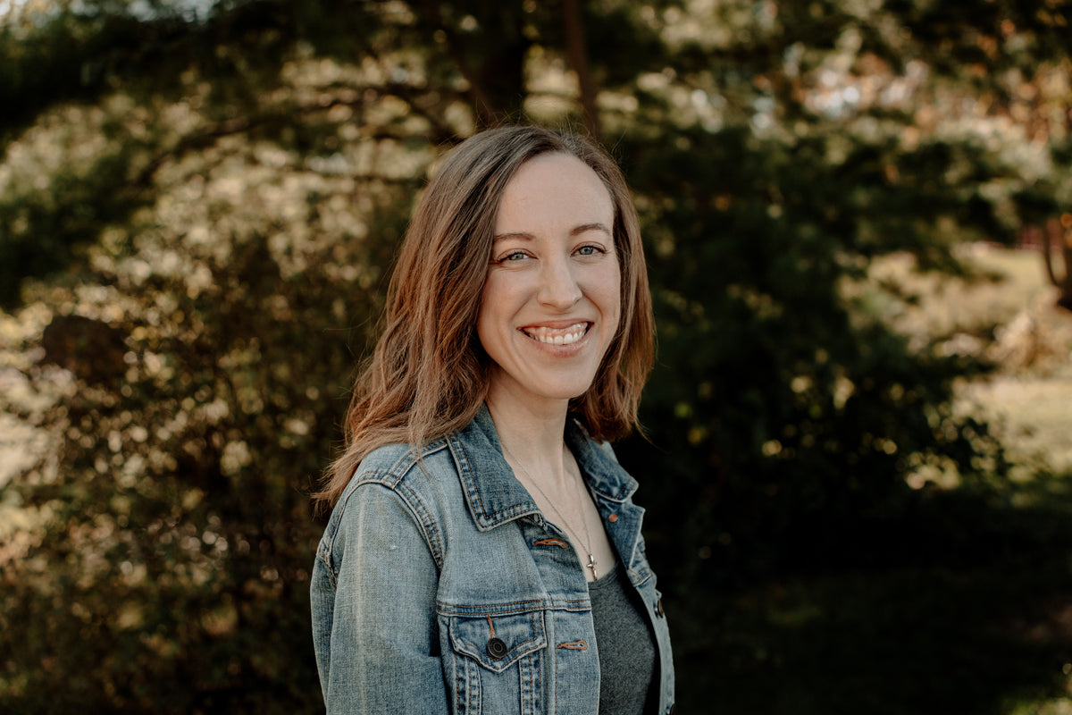 A smiling woman with long brown hair wearing a light-wash denim jacket over a dark top, photographed outdoors against a softly blurred background of trees and foliage.