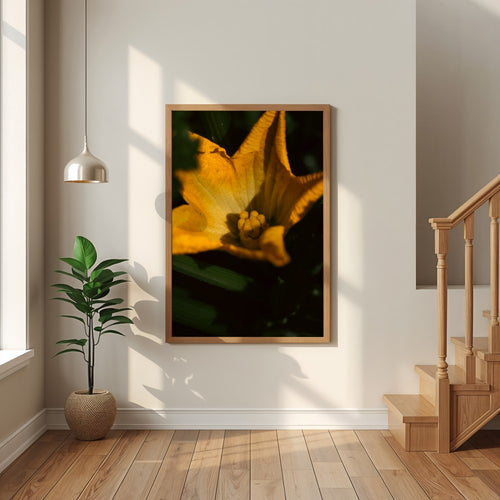 A framed vertical art print of a close-up yellow flower with dark green and black shadows, displayed on a white wall in a modern home interior with a wooden staircase, potted plant, and pendant light.
