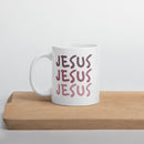 White mug with 'JESUS JESUS JESUS' printed in pink on a wooden surface with a light gray background