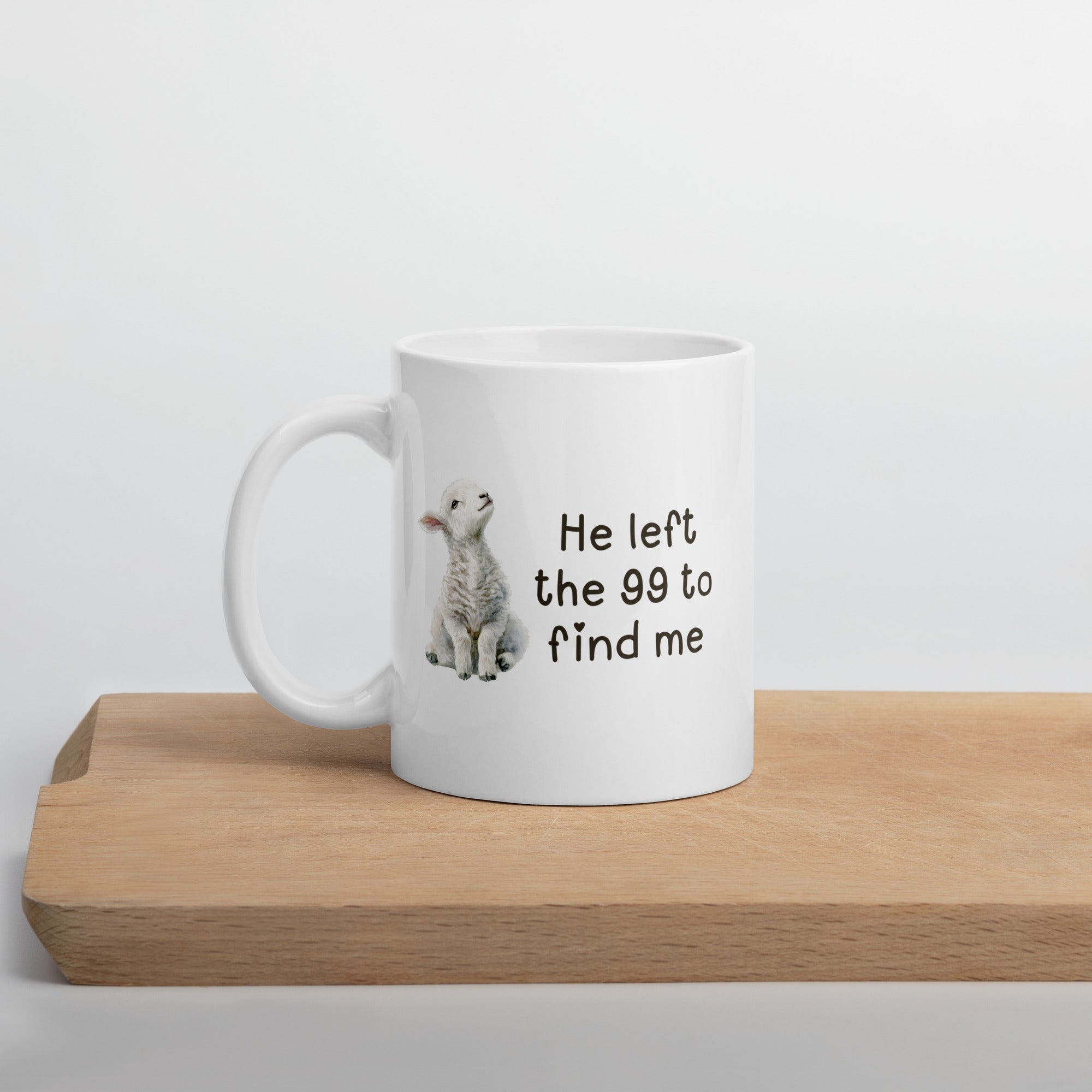 white 11 oz mug with a watercolor lamb and 'He left the 99 to find me' text sitting on a wooden board
